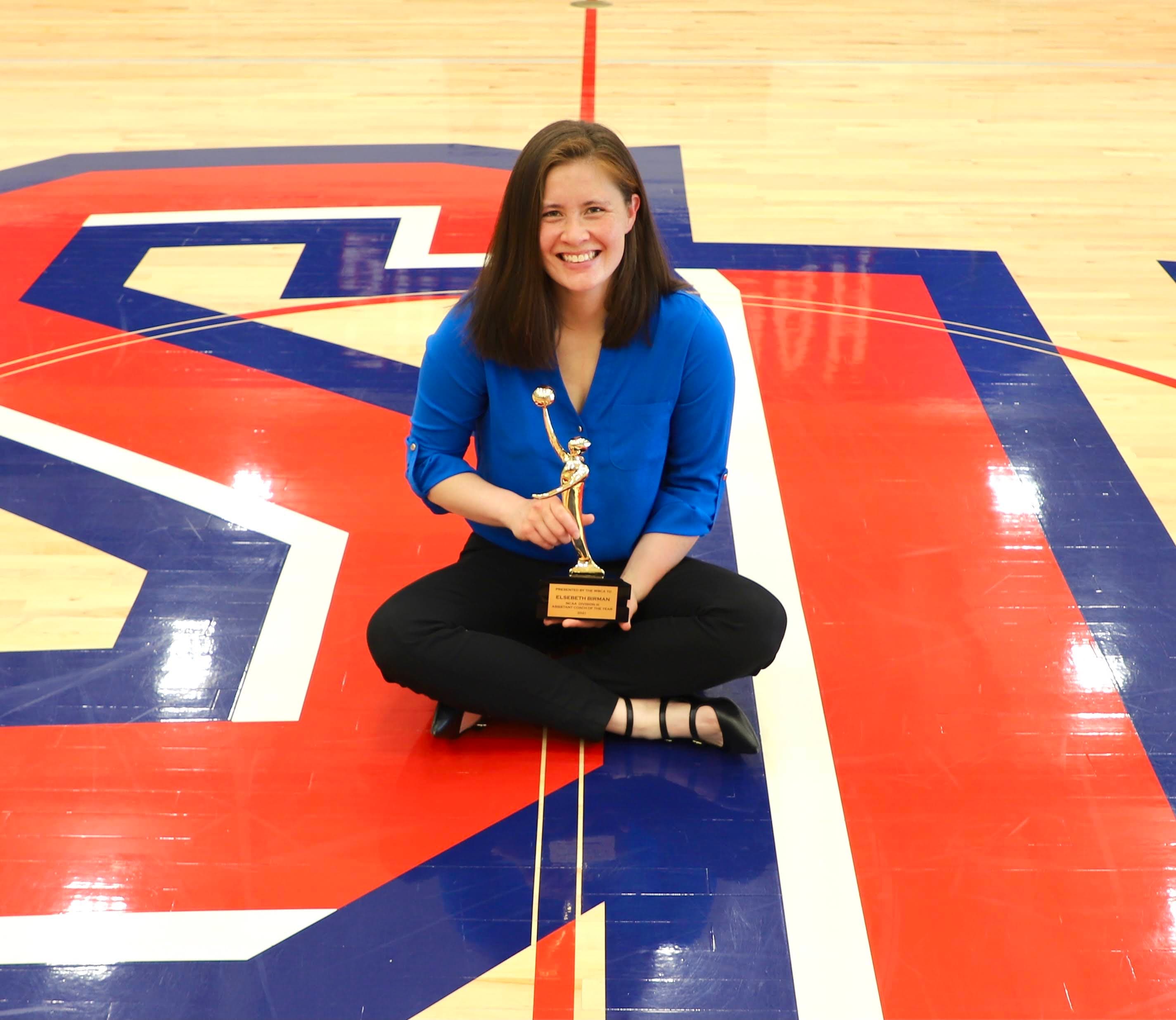 Coach El seated on the court at Shenandoah University with her WBCA award.